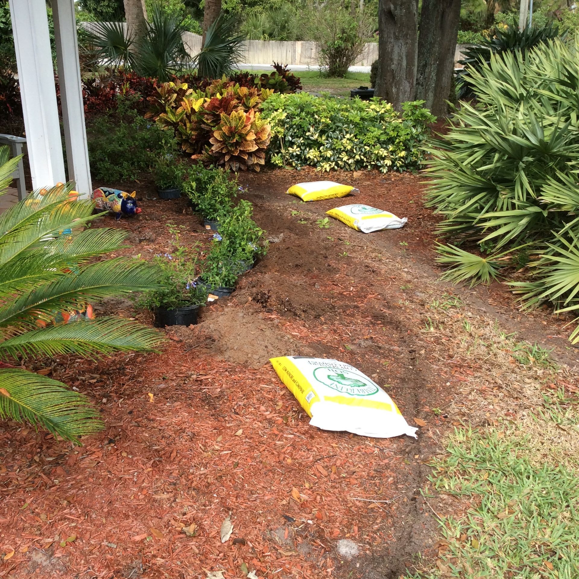 Garden area with various plants and bushes being planted, surrounded by bags of fertilizer and mulch.